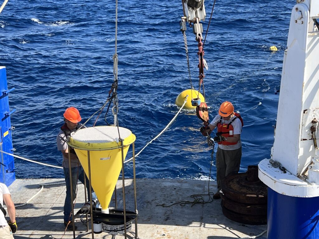 Two scientists prepare a conical sediment trap on the aft deck of a ship. A tethered buoy floats in the water in the background.